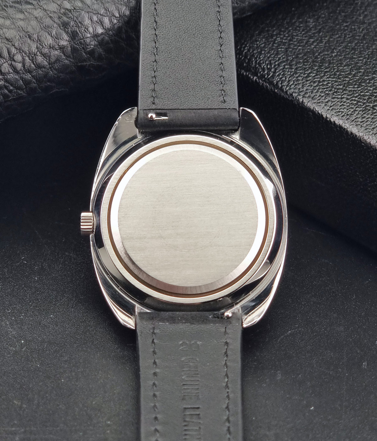 Close-up of the back of a wristwatch with a brushed silver-tone case and a black leather strap on a dark textured background.