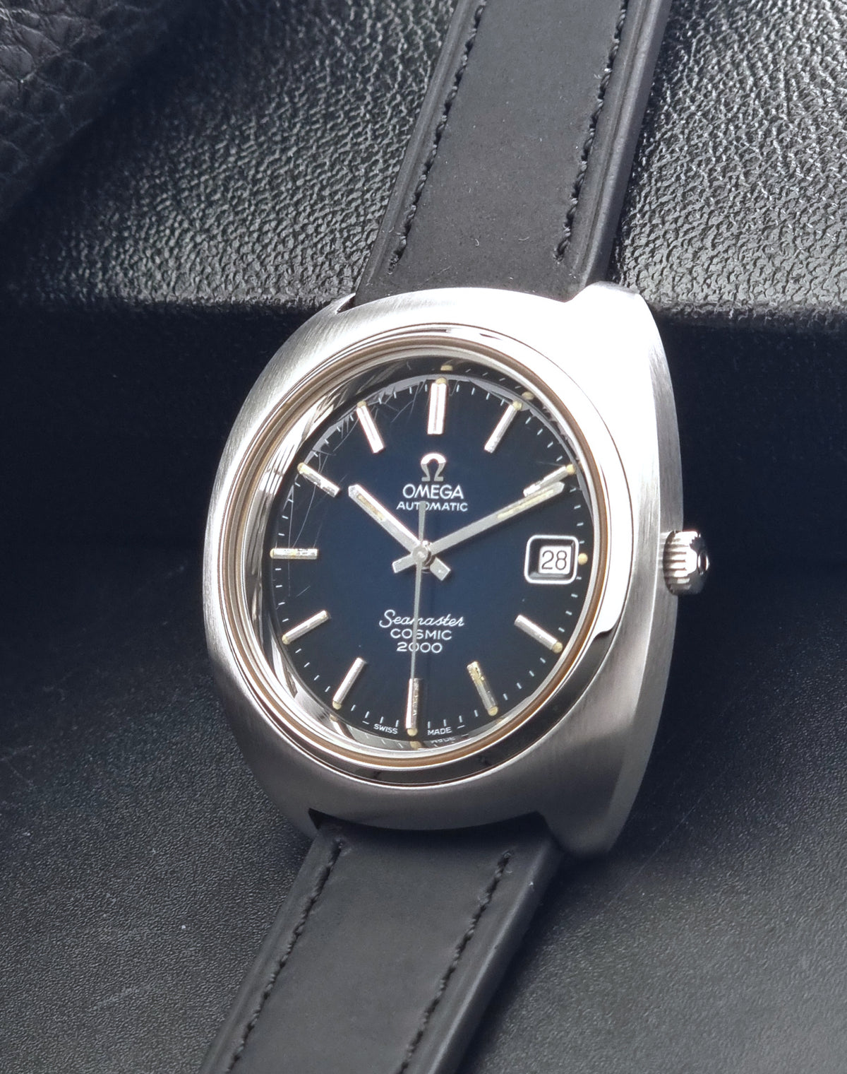 Vintage Omega Wristwatch 166.132 Seamaster Cosmic 2000 with a dark blue dial, silver-tone stainless steel case, and a black leather strap, resting on a textured black surface.