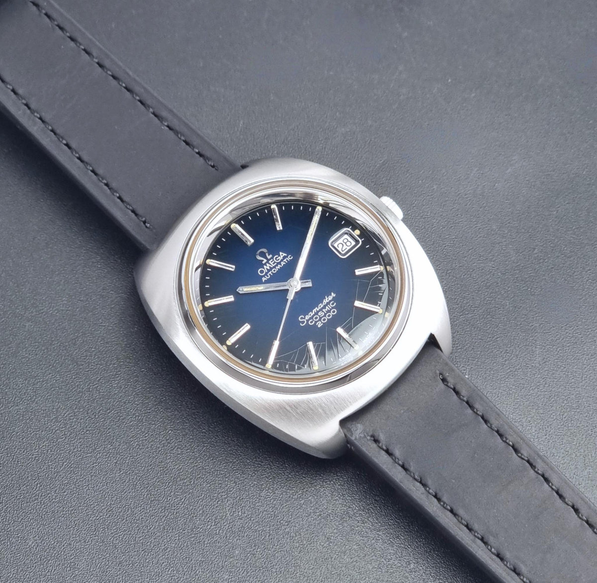 Vintage Omega Wristwatch 166.132 Seamaster Cosmic 2000 with a brushed stainless steel case, a deep blue dial, and a dark gray leather strap, shown on a textured gray background.