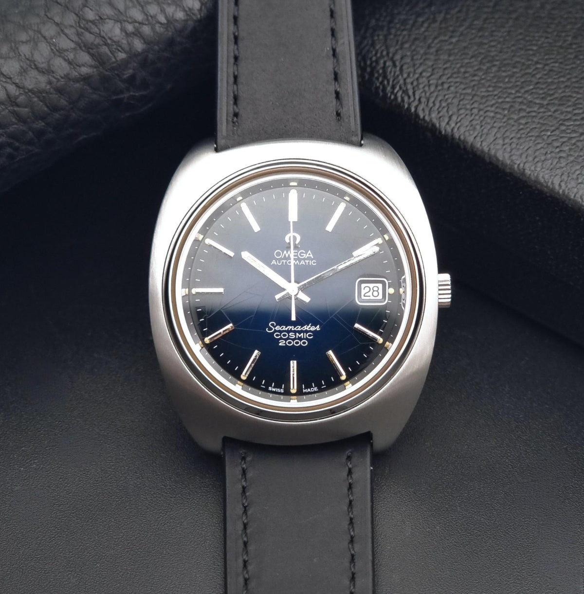 Vintage Omega Wristwatch 166.132 Seamaster Cosmic 2000 with a silver-toned stainless steel case, dark blue dial, and black leather strap, displayed on a textured black leather surface.