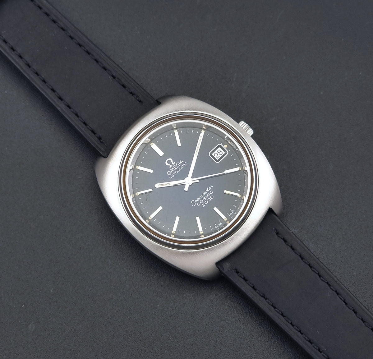 Vintage Omega Wristwatch 166.132 Seamaster Cosmic 2000  with a dark blue dial, silver-tone stainless steel case, and a black leather strap, shown diagonally on a dark gray textured background.