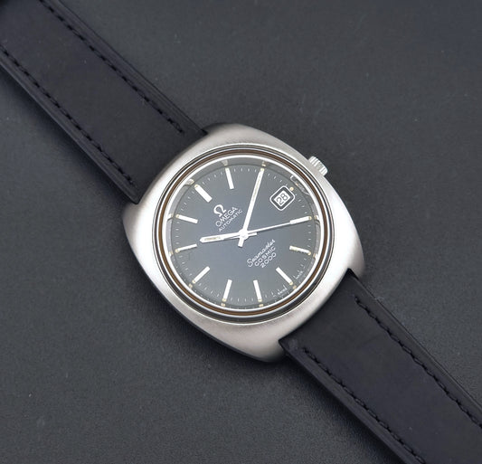 Vintage Omega Wristwatch 166.132 Seamaster Cosmic 2000  with a dark blue dial, silver-tone stainless steel case, and a black leather strap, shown diagonally on a dark gray textured background.