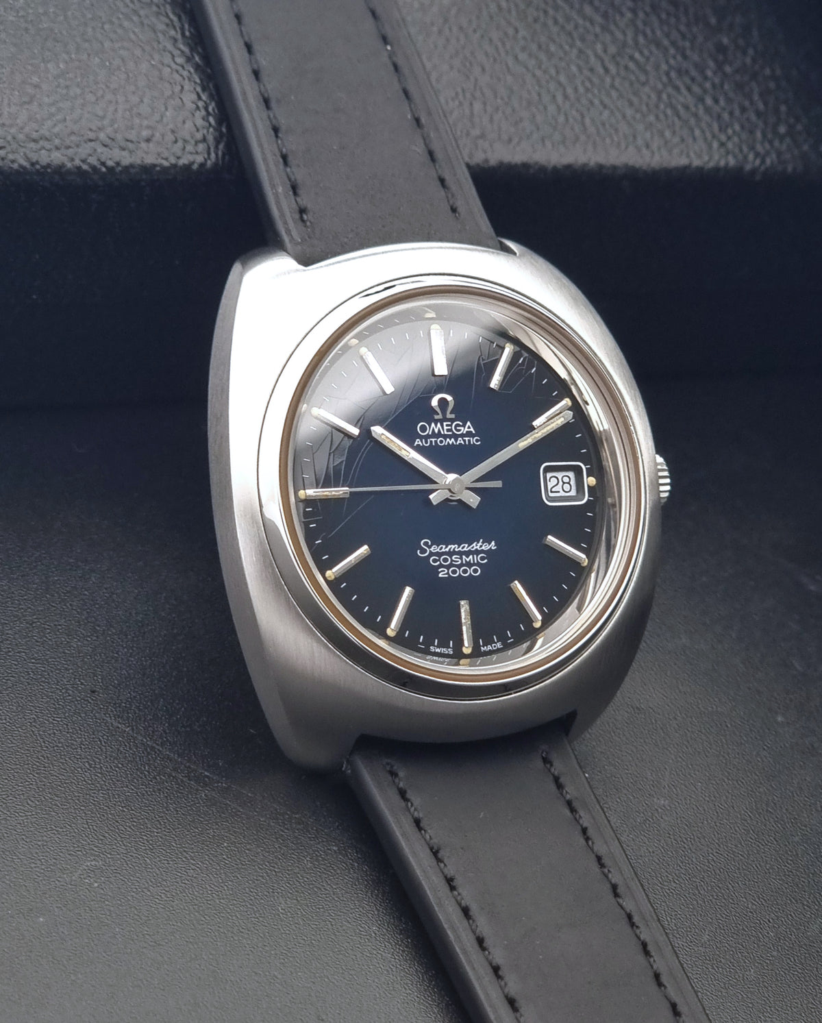 Vintage Omega Wristwatch 166.132 Seamaster Cosmic 2000 with a silver-tone case, dark blue dial, and black leather strap, displayed on a textured black surface.
