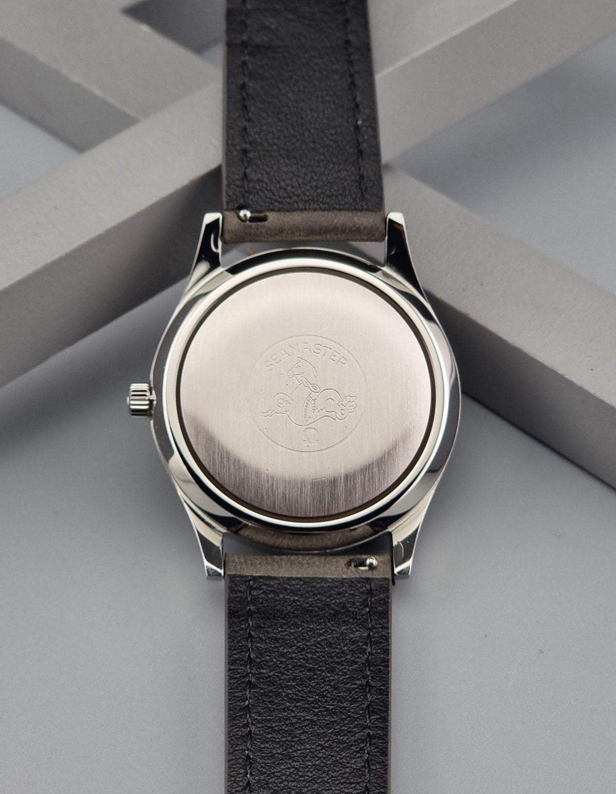 Close-up of the back of a wristwatch with a brushed silver-tone round case and a dark gray textured strap, photographed on a light gray geometric surface.