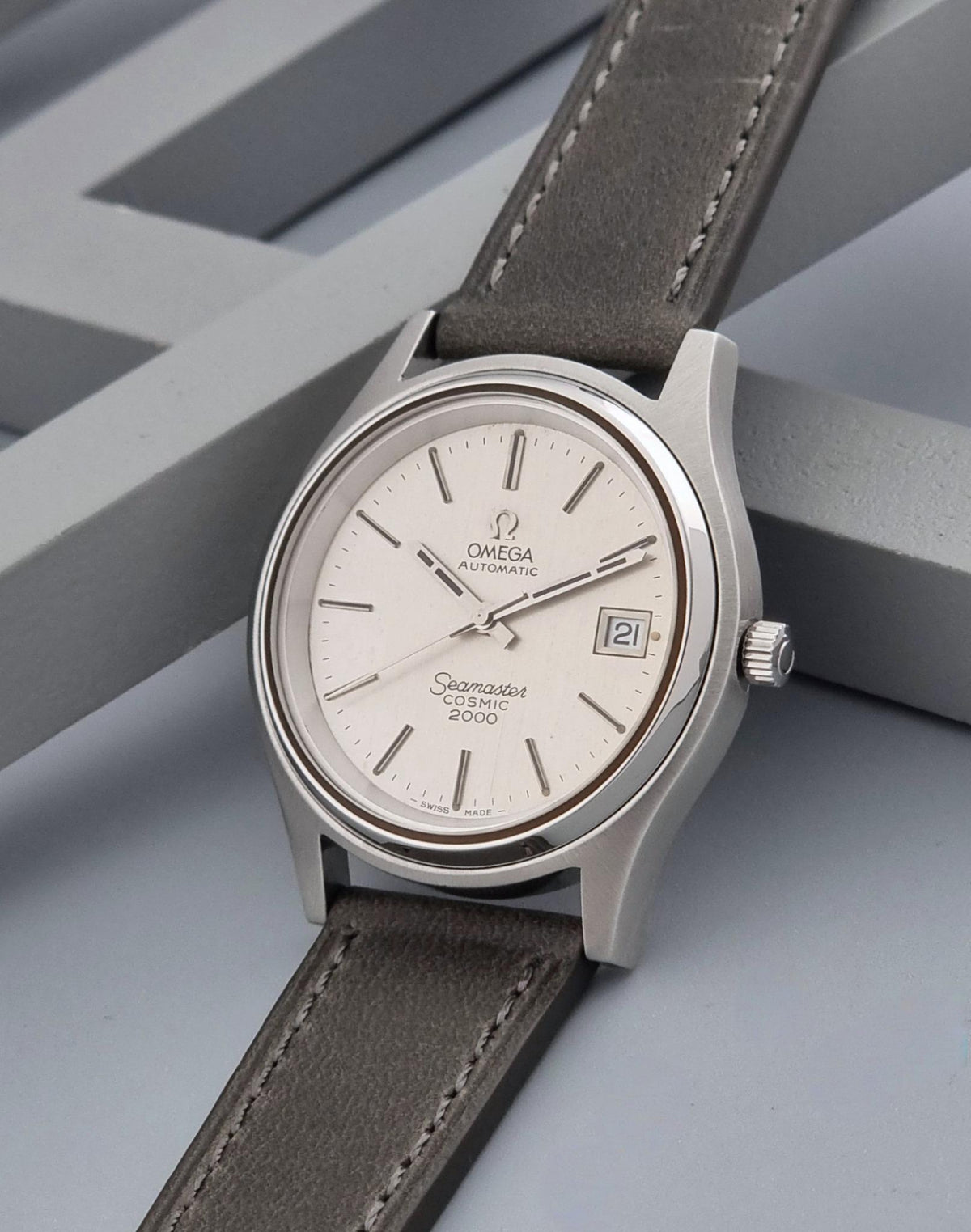 Vintage Omega Wristwatch 166.128 Seamaster Cosmic 2000 with a silver stainless steel case, cream dial, and a dark gray leather strap, resting on a gray geometric background.