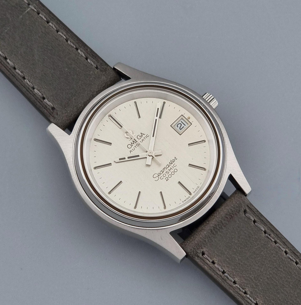 Vintage Omega Wristwatch 166.128 Seamaster Cosmic 2000 with a silver stainless steel case, white dial, and a dark gray leather strap, shown on a neutral gray background.