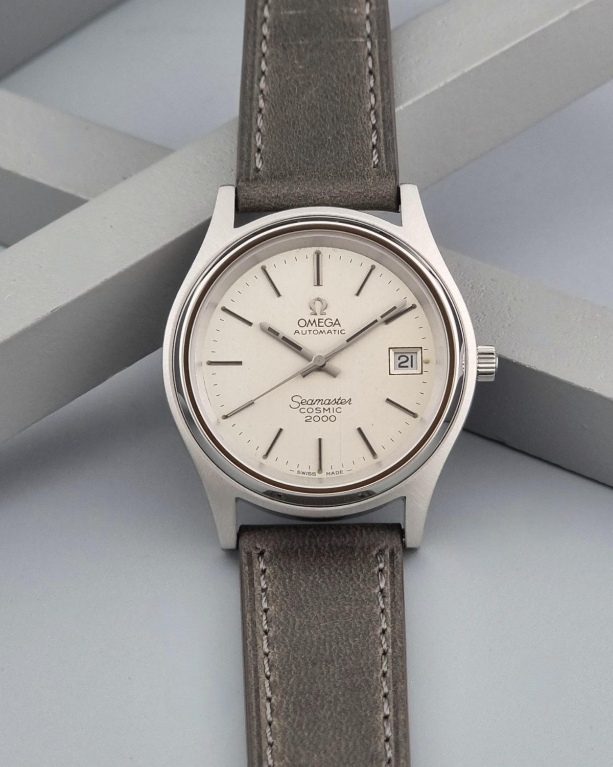Vintage Omega Wristwatch 166.128 Seamaster Cosmic 2000 with a silver stainless steel case and a taupe-brown leather strap, displayed on a light gray geometric background.