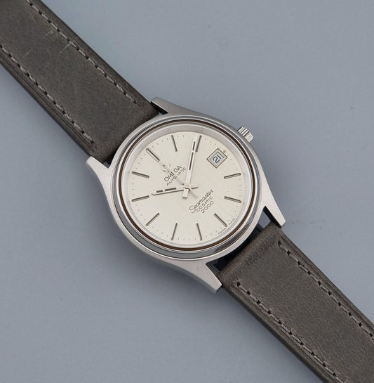 Vintage Omega Wristwatch 166.128 Seamaster Cosmic 2000 with a silver-tone stainless steel case, white dial, and a dark gray leather strap, shown on a solid gray background.