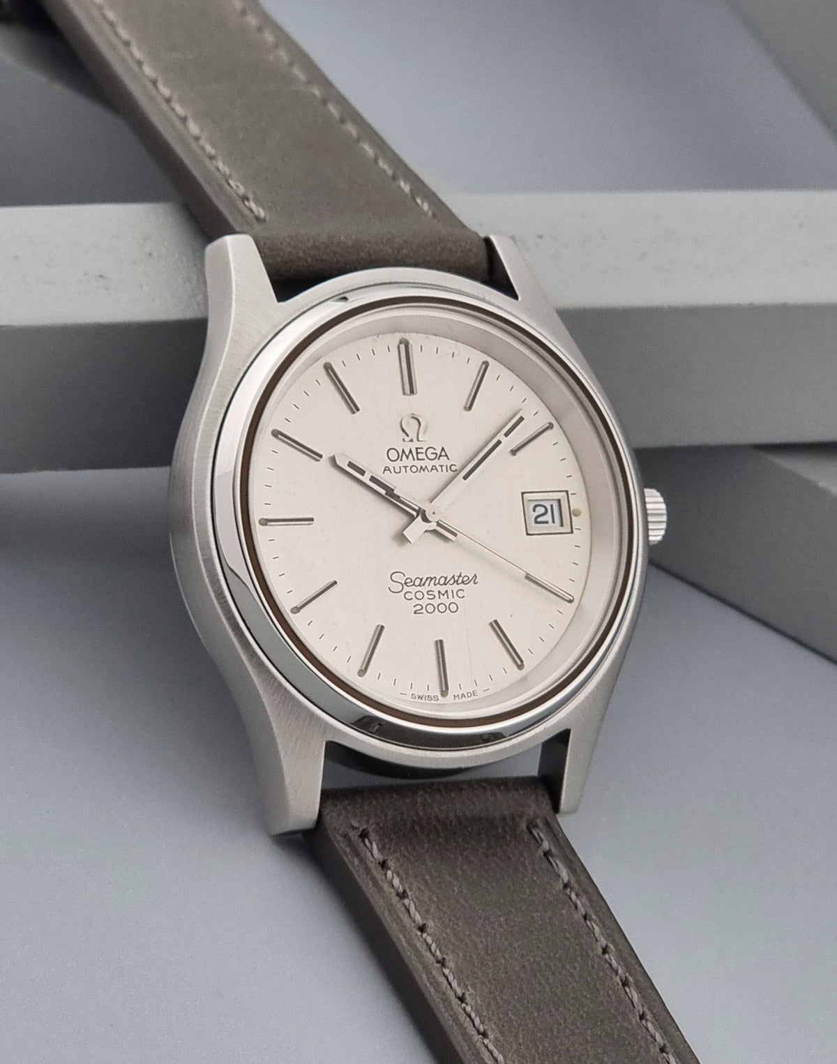 An Omega Seamaster 2000 watch with a silver stainless steel case, a white dial, and a taupe-brown leather strap, shown resting on a gray geometric surface.