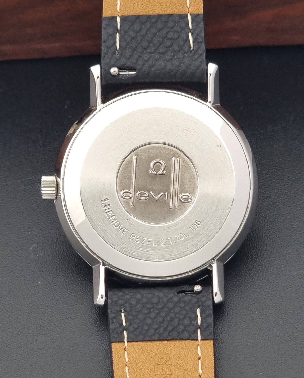 Close-up of the back of a Vintage Omega  De-Ville 166.0161 watch with a silver-tone round case and a dark textured strap featuring a leather panels and white stitching. The case is engraved with the Deville brand name and logo.