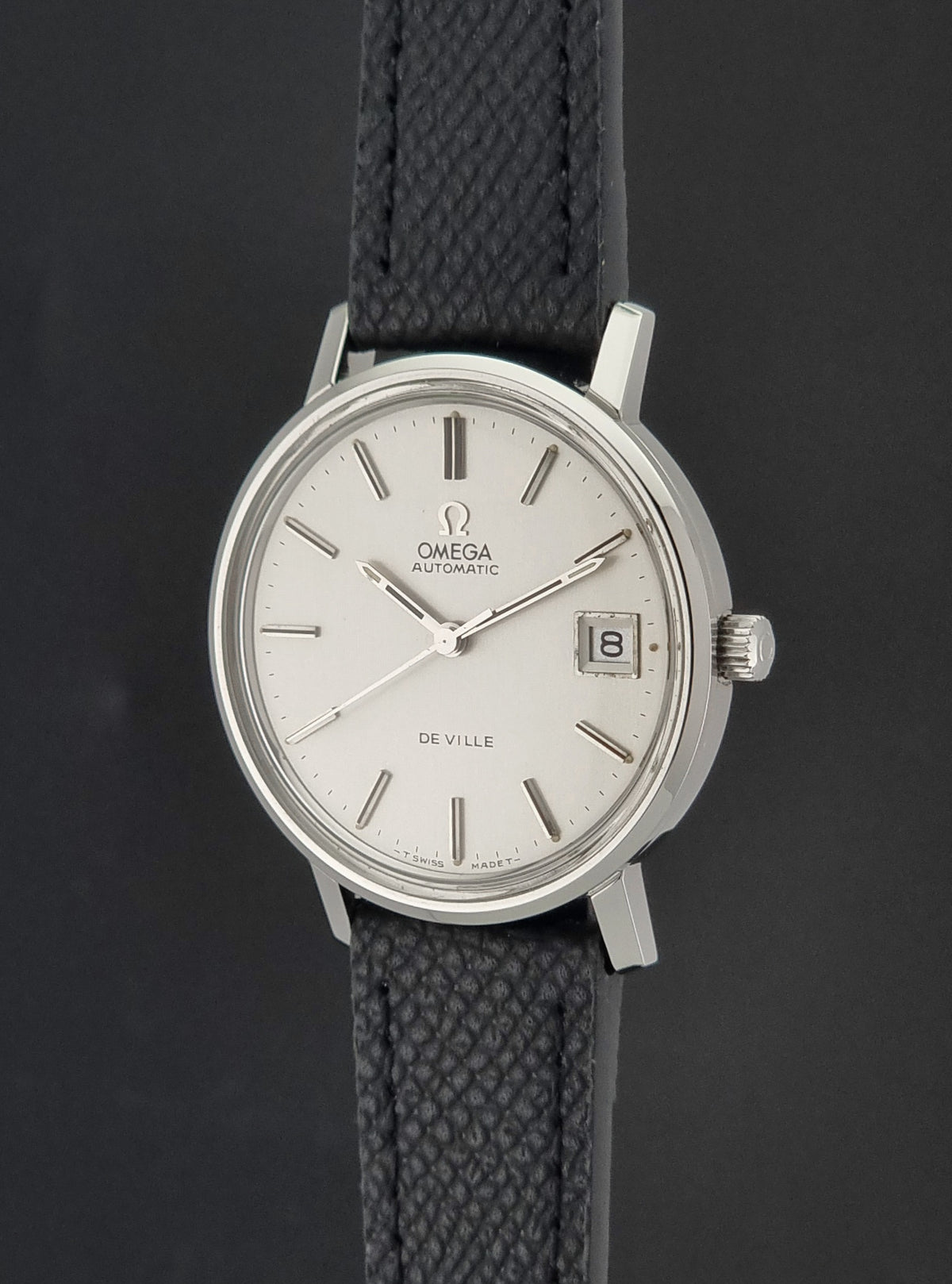 An Omega De Ville wristwatch with a silver-toned oval case, white dial, and textured black strap, shown against a dark gray background.