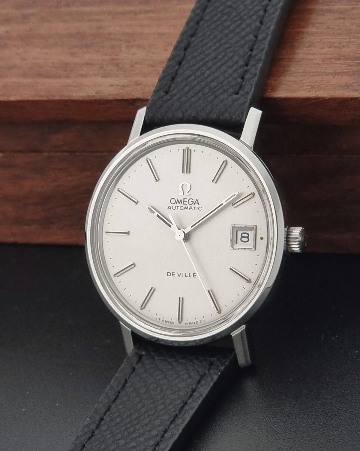 Omega De Ville Automatic wristwatch with a silver-toned oval case, white dial, and black textured leather strap, resting on a dark surface with a wooden background.