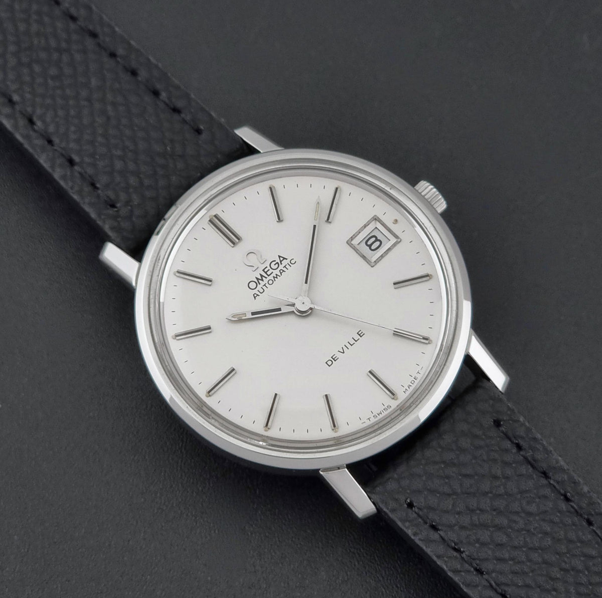 A vintage Omega De Ville automatic watch with a silver-toned oval case, white dial, and black textured leather strap, shown on a dark gray background.
