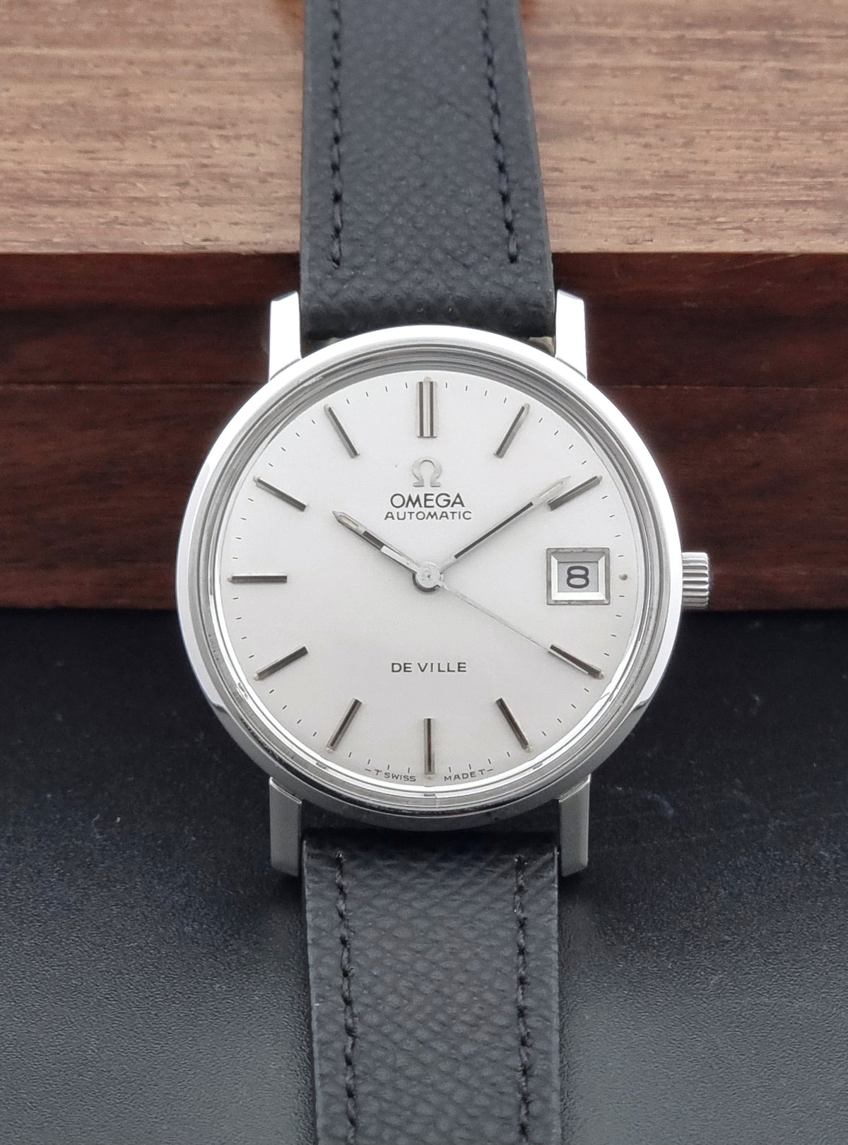 An Omega De Ville automatic wristwatch with a silver-toned round case, white dial, and a dark gray textured leather strap, displayed against a dark background with a wooden block behind it.