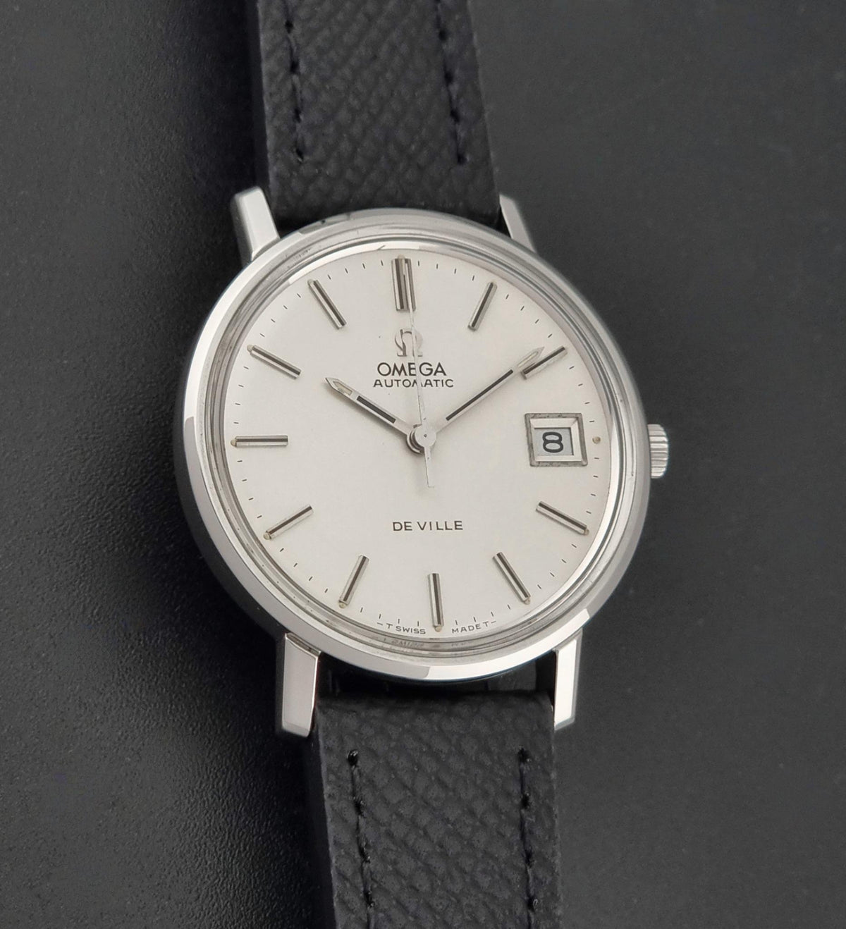 Omega De Ville Automatic wristwatch with a silver stainless steel case, white dial, and a textured black leather strap, shown on a dark gray background.