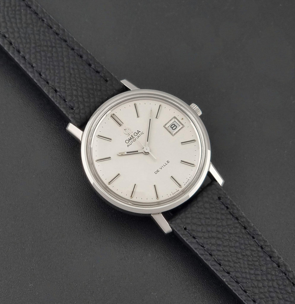 An Omega Deville wristwatch with a silver-toned metal case, white dial, and a textured black leather strap, shown on a dark gray background.