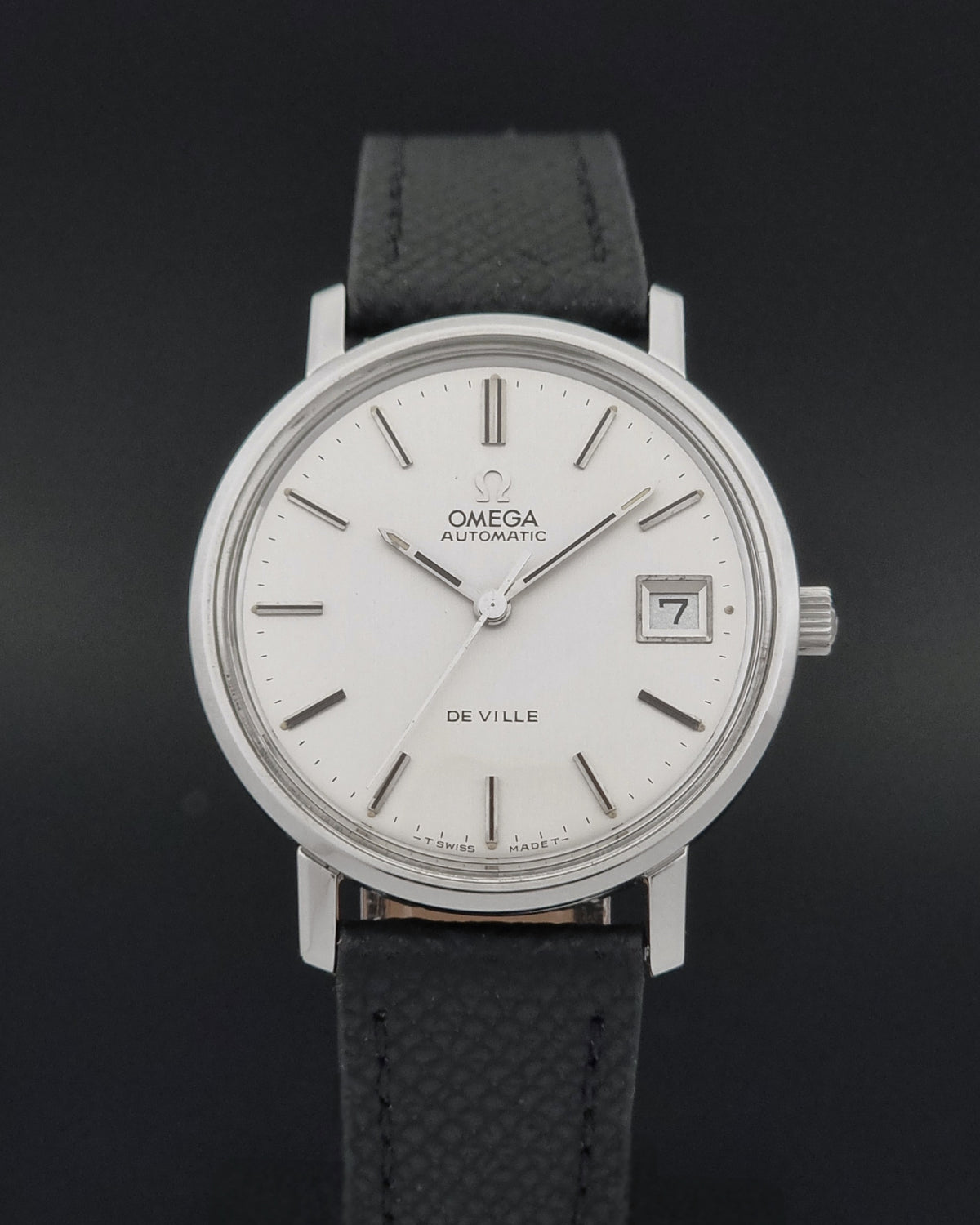 Omega De Ville Automatic watch with a silver-toned round case, white dial, and black textured leather strap, shown against a dark background.