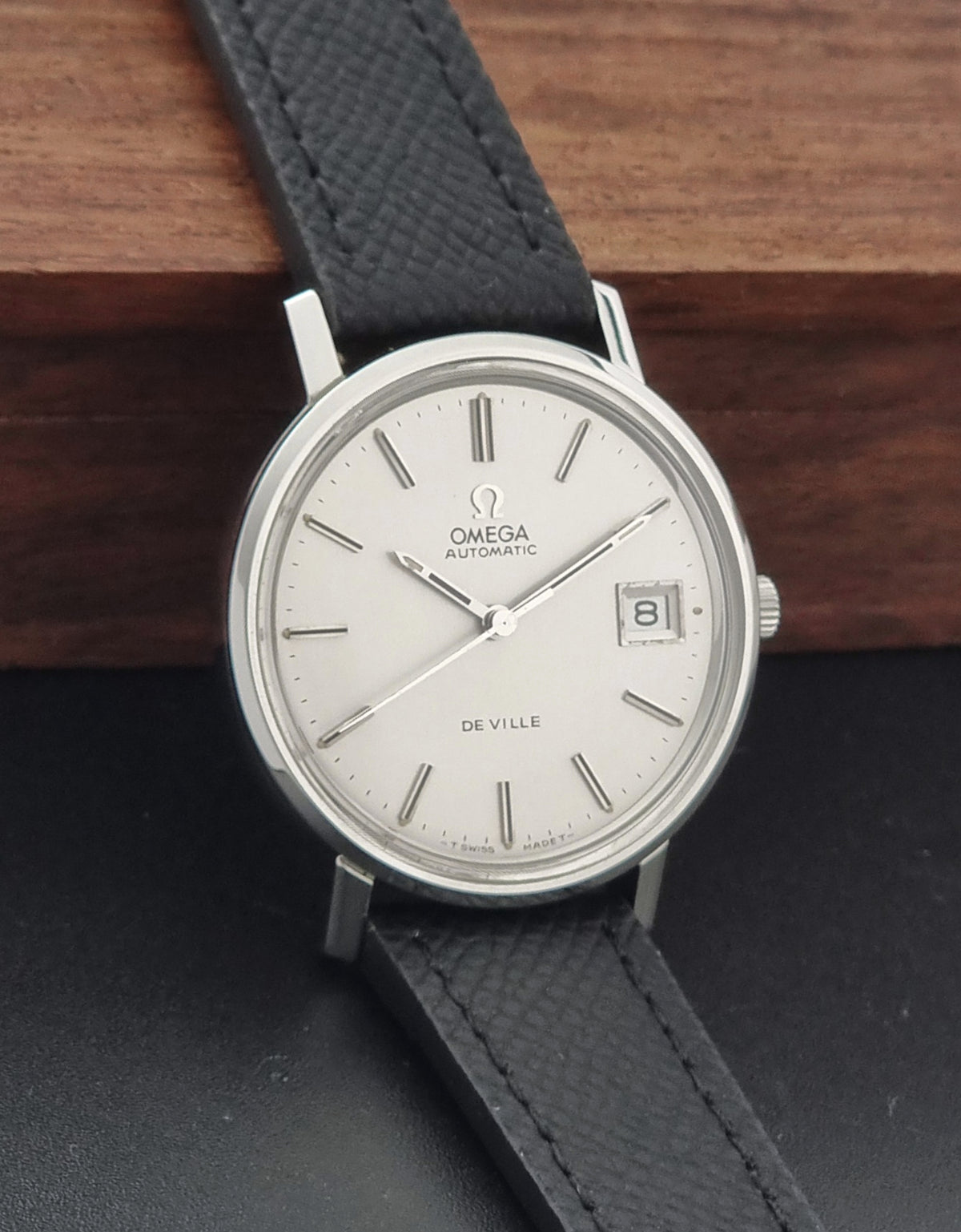 An Omega De Ville Automatic wristwatch with a silver-toned oval case, white dial, and textured black leather strap, resting on a dark surface with a wooden background.