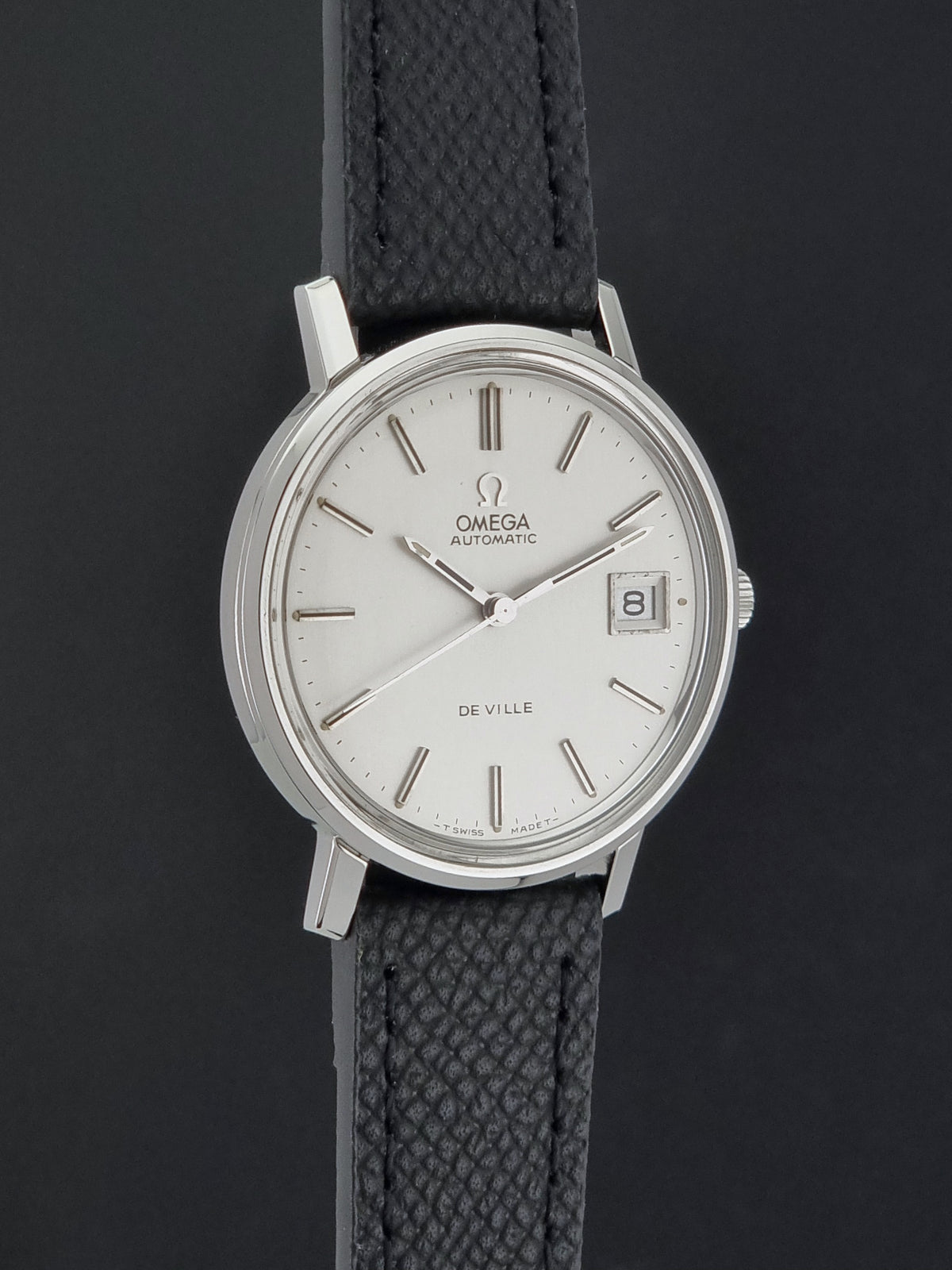 Omega De Ville Automatic wristwatch with a silver-toned oval case, white dial, and textured black strap, shown against a dark gray background.