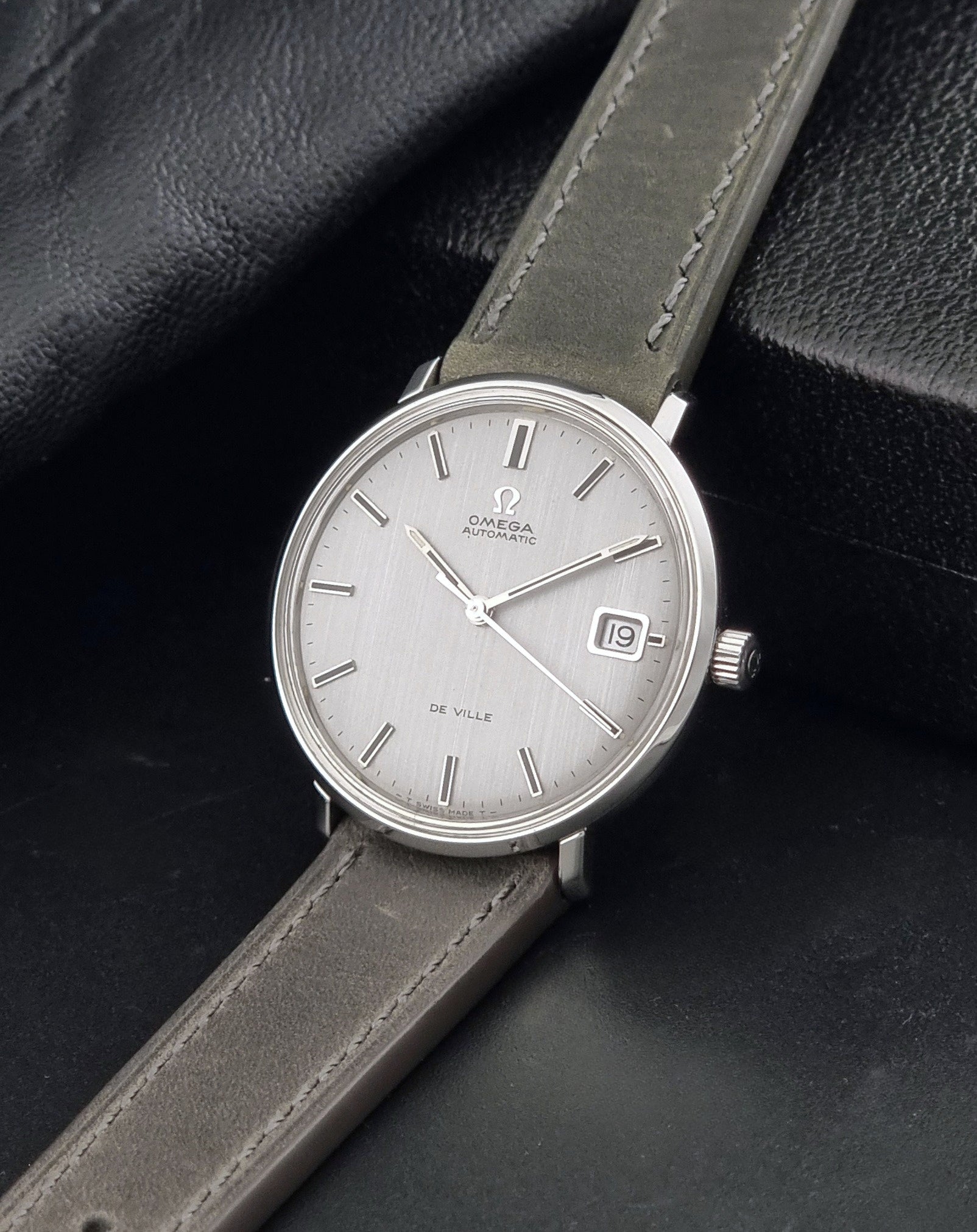 Vintage Omega De-Ville 166.033 Wristwatch with a date window, attached to a gray leather strap, photographed on a dark gray surface crown view