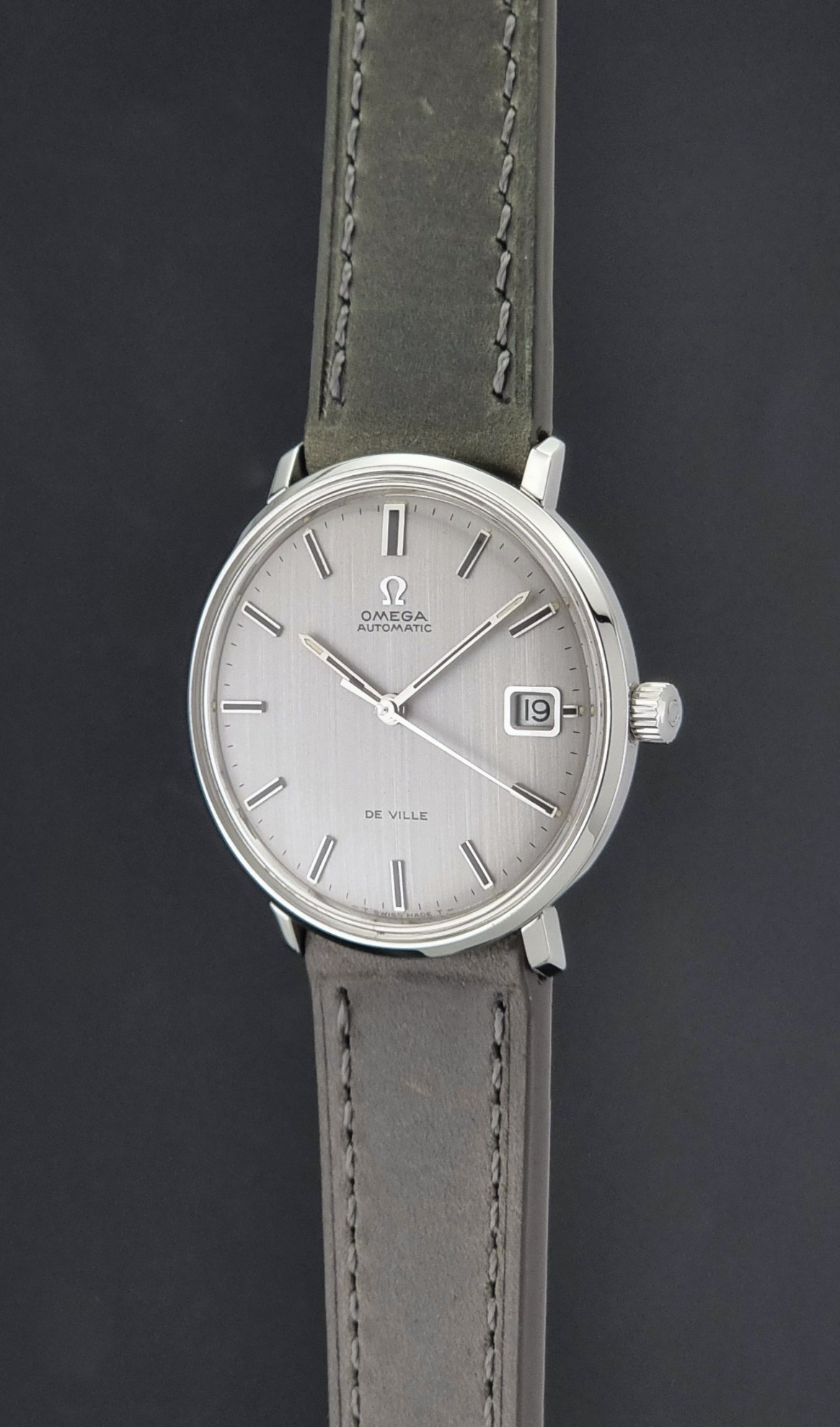 Vintage Omega De-Ville 166.033 Wristwatch with a date window, attached to a gray leather strap, photographed on a dark gray surface crown profile view