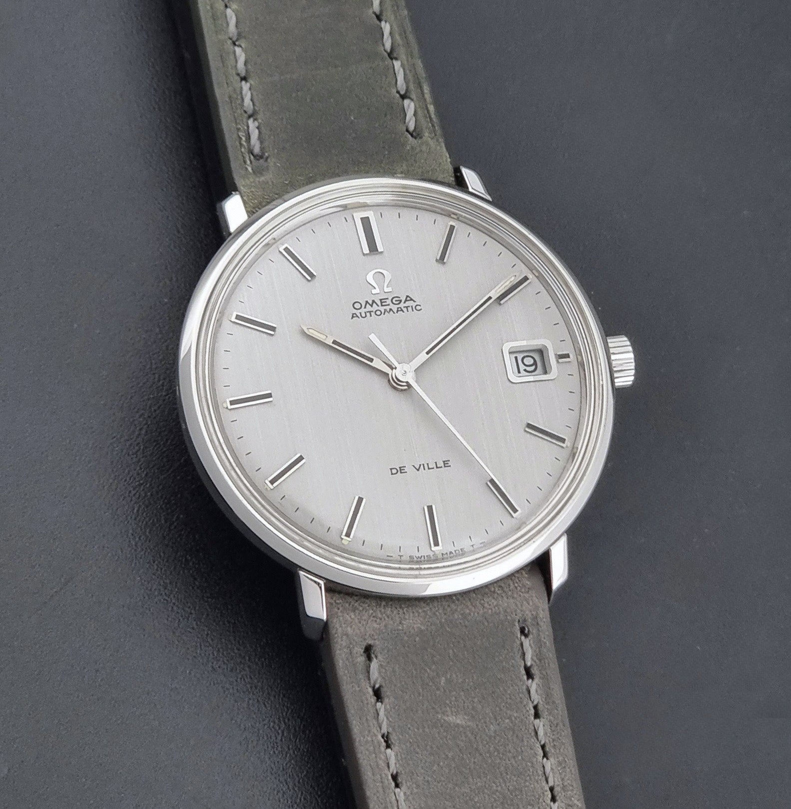Vintage Omega De-Ville 166.033 Wristwatch with a date window, attached to a gray leather strap, photographed on a dark gray surface front view