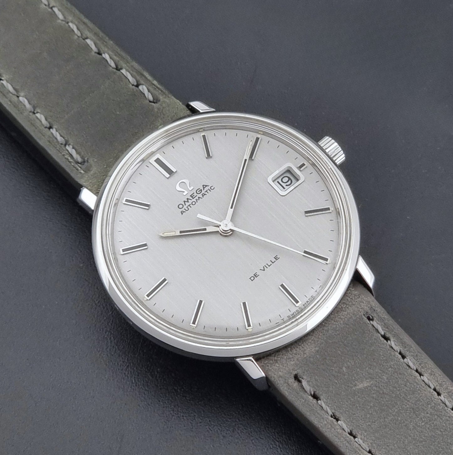 Vintage Omega De-Ville 166.033 Wristwatch with a date window, attached to a gray leather strap, photographed on a dark gray surface close dial view