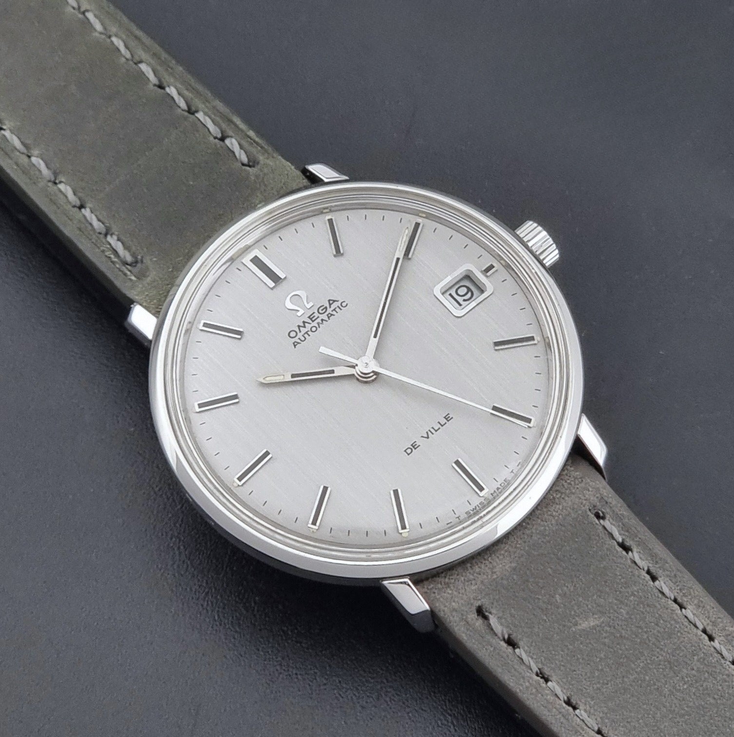 Vintage Omega De-Ville 166.033 Wristwatch with a date window, attached to a gray leather strap, photographed on a dark gray surface close dial view