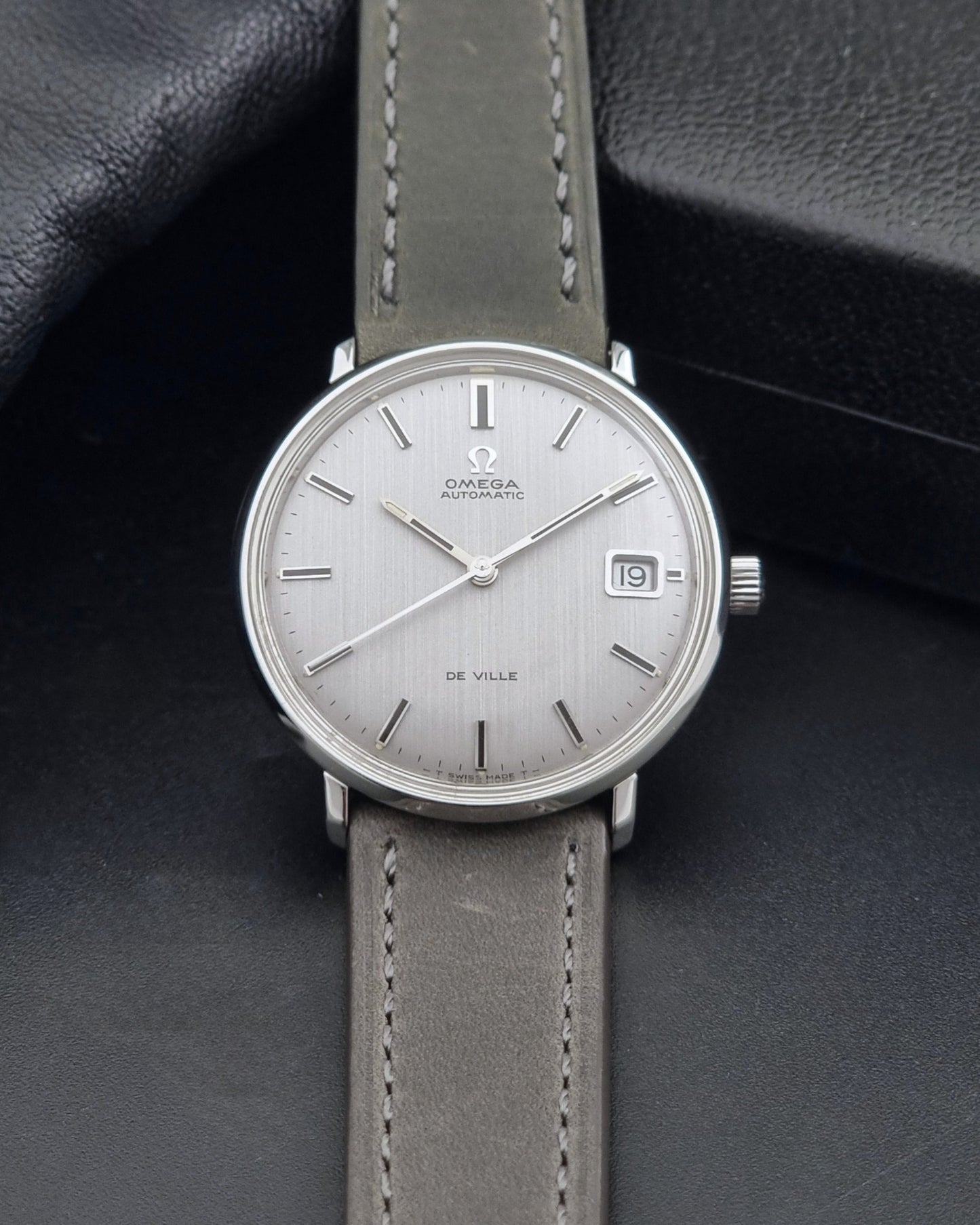 Vintage Omega De-Ville 166.033 Wristwatch with a date window, attached to a gray leather strap, photographed on a dark gray surface dial view