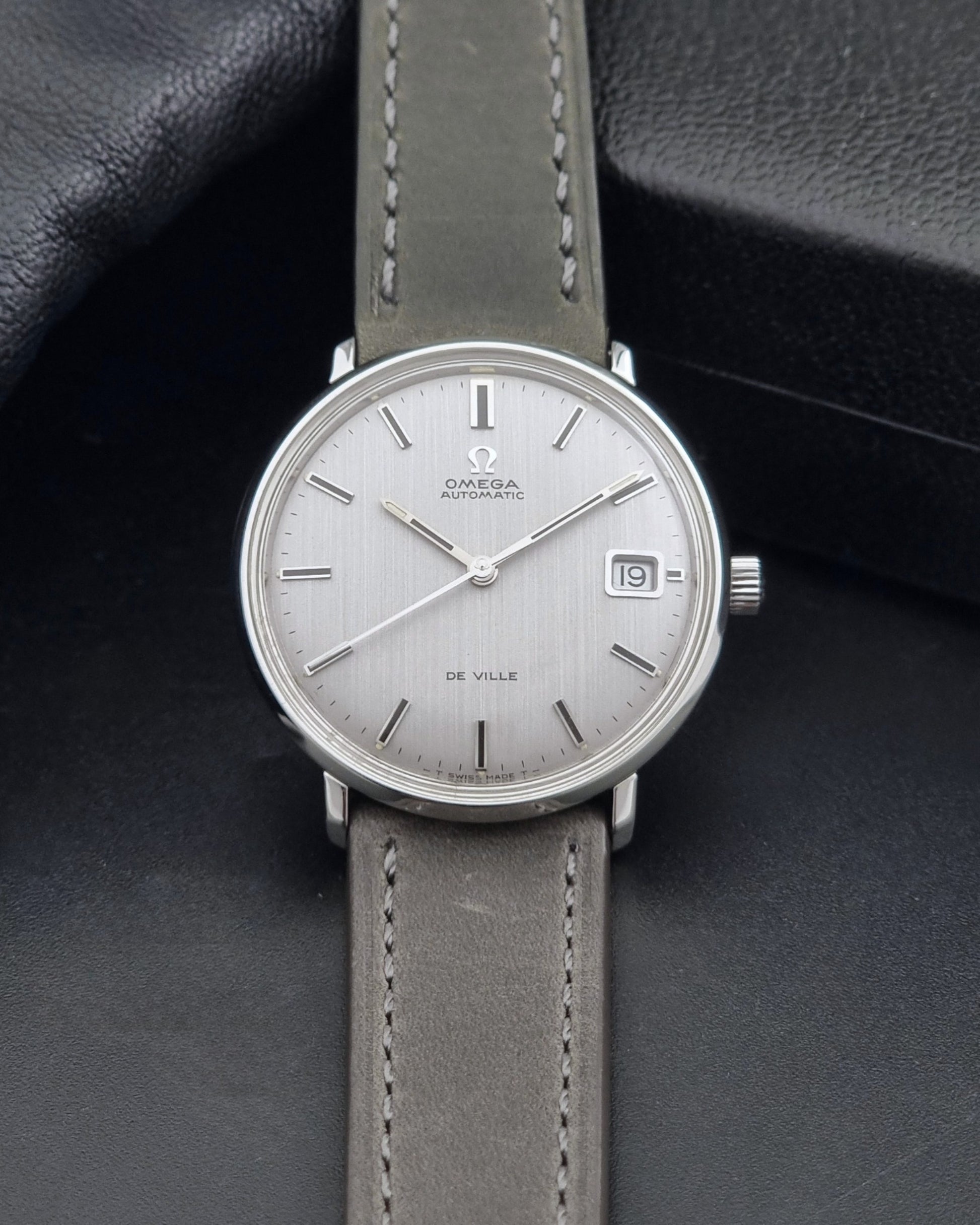Vintage Omega De-Ville 166.033 Wristwatch with a date window, attached to a gray leather strap, photographed on a dark gray surface dial view