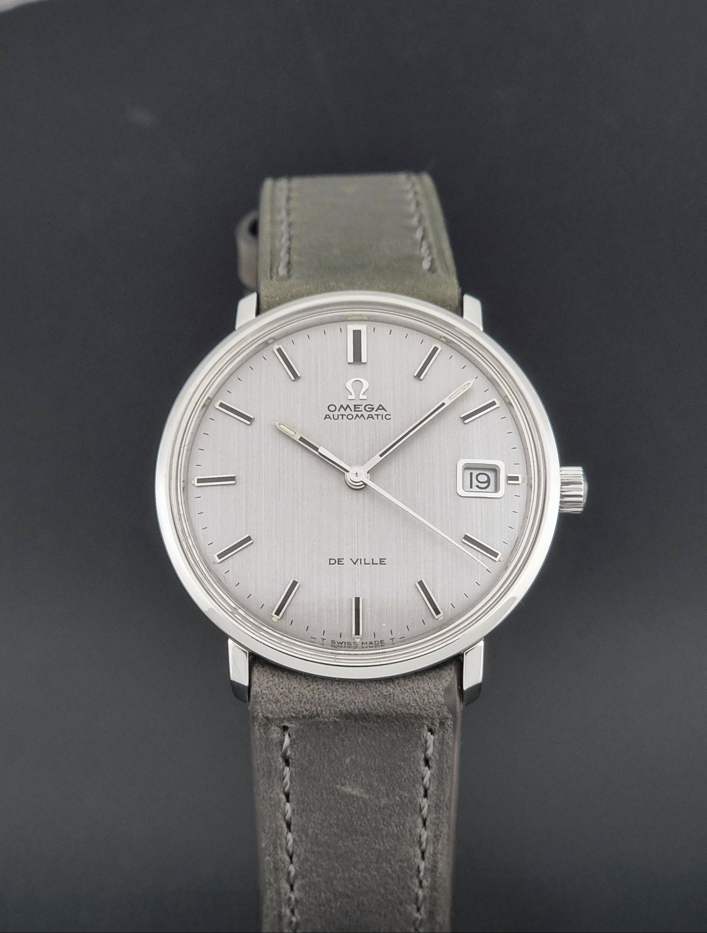 Vintage Omega De-Ville 166.033 Wristwatch with a date window, attached to a gray leather strap, photographed on a dark gray surface close up dial view
