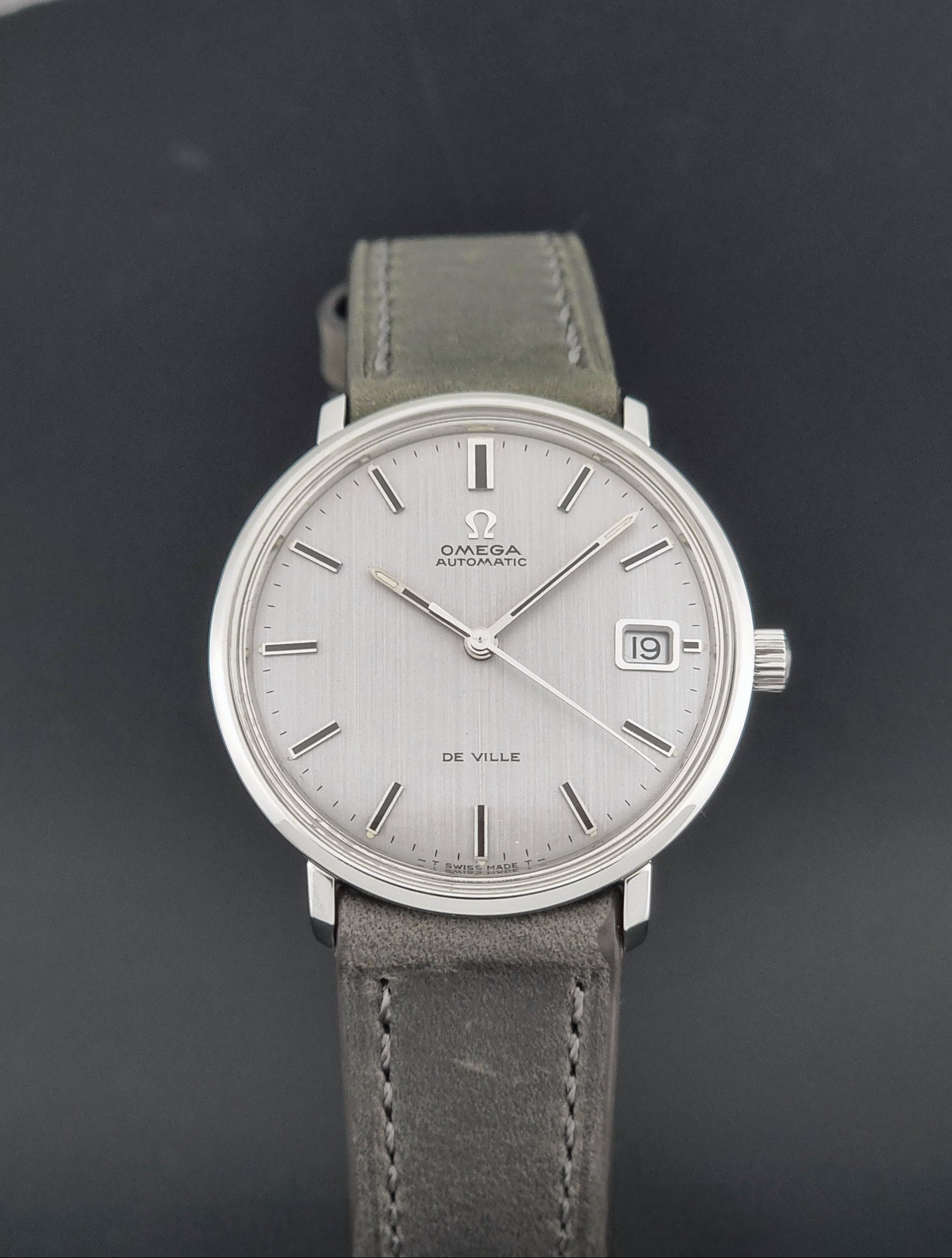 Vintage Omega De-Ville 166.033 Wristwatch with a date window, attached to a gray leather strap, photographed on a dark gray surface close up dial view