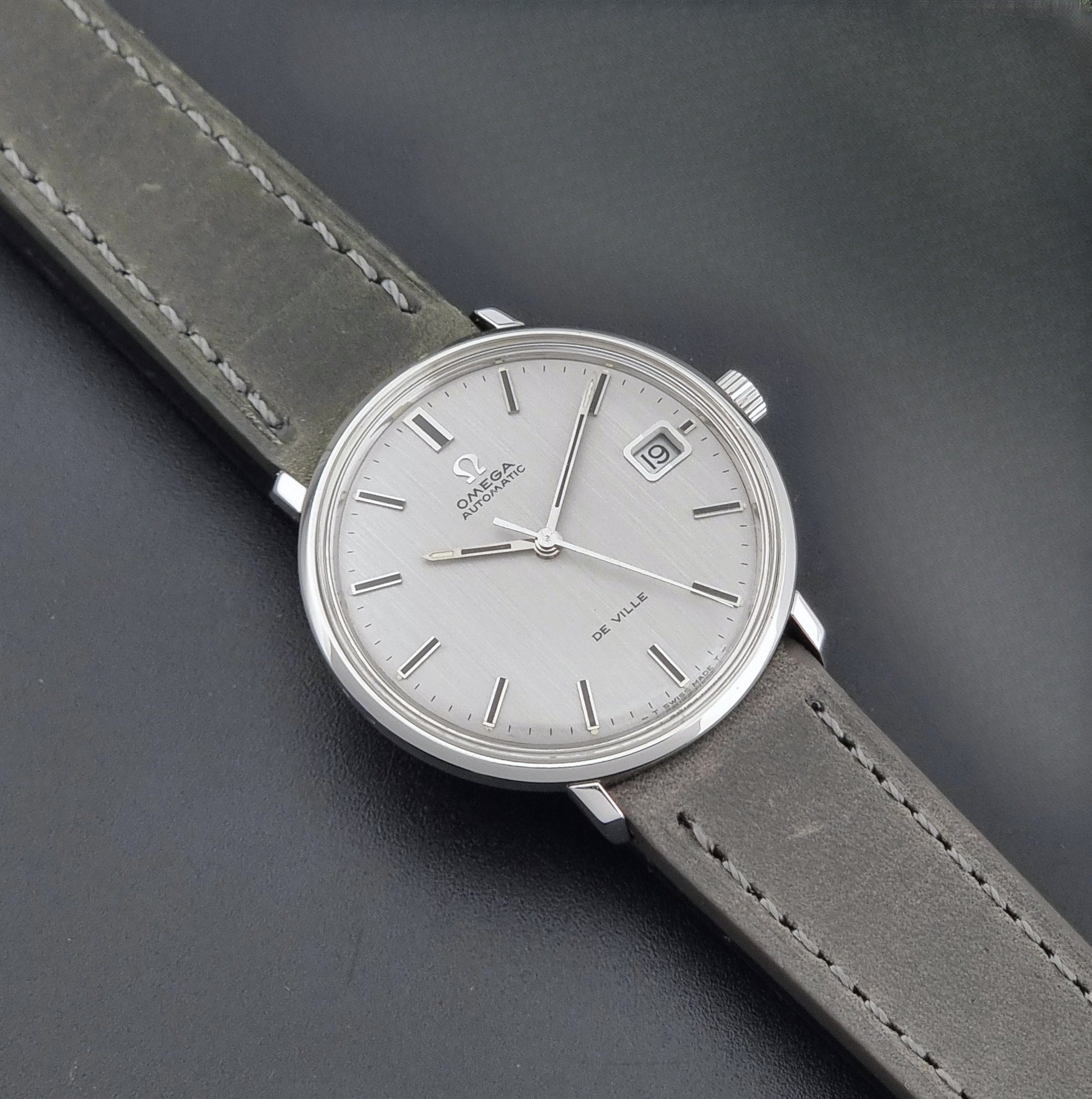 Vintage Omega De-Ville 166.033 Wristwatch with a date window, attached to a gray leather strap, photographed on a dark gray surface dial