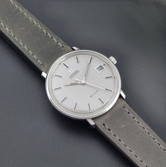 Vintage Omega De-Ville 166.033 Wristwatch with a date window, attached to a gray leather strap, photographed on a dark gray surface dial