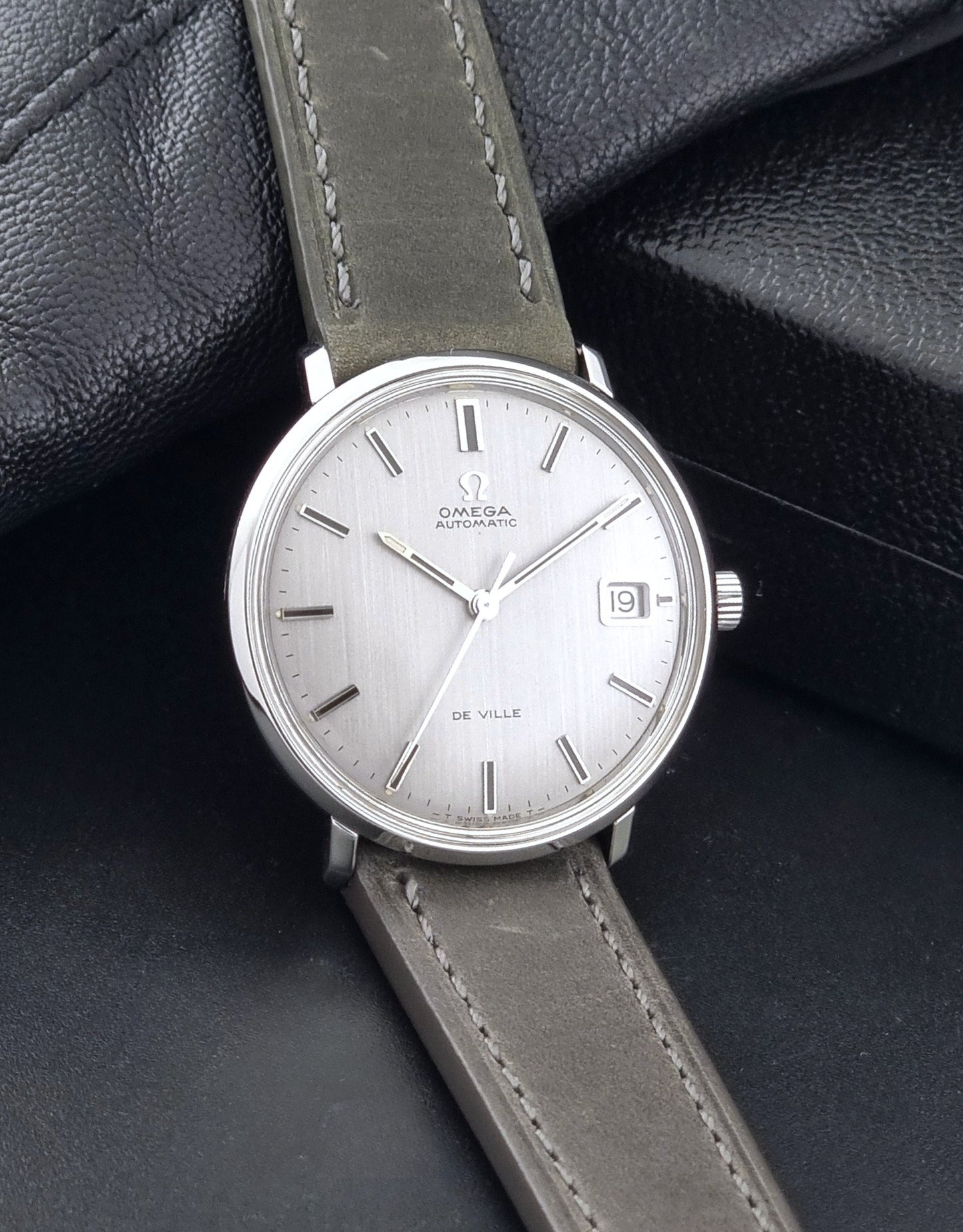 Vintage Omega De-Ville 166.033 Wristwatch with a date window, attached to a gray leather strap, photographed on a dark gray surface side view