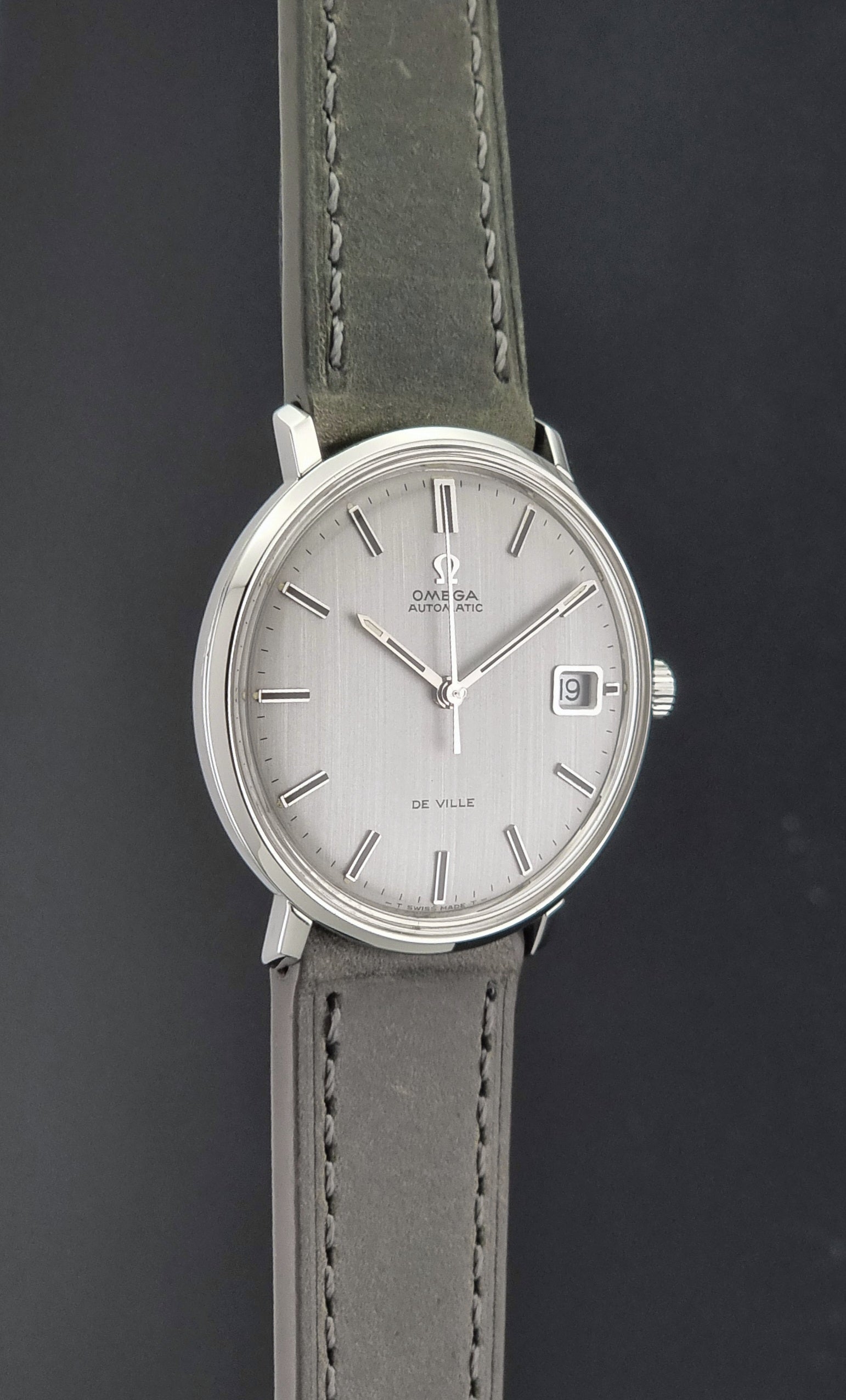 Vintage Omega De-Ville 166.033 Wristwatch with a date window, attached to a gray leather strap, photographed on a dark gray surface side profile view