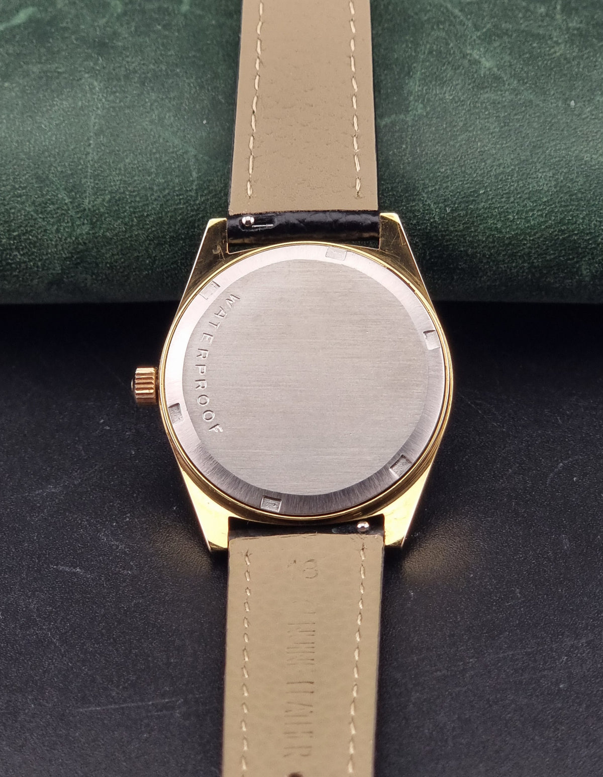 Close-up of the back of a Vintage Omega Geneve 135.041 watch with a round brushed-metal case and a light beige leather strap, resting on a dark surface with a green textured background.