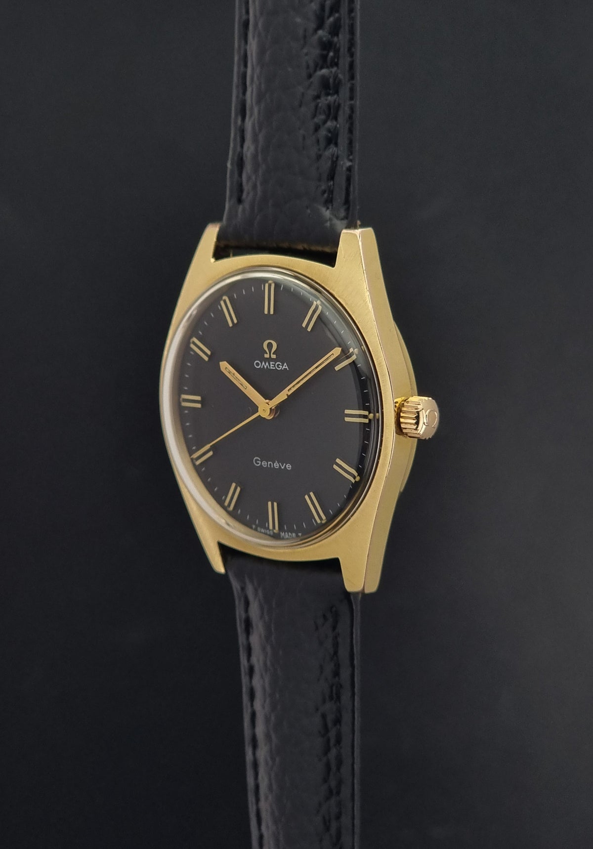 Vintage Omega Geneve 135.041 watch with a gold-tone case, black dial, and black leather strap, shown against a dark background.