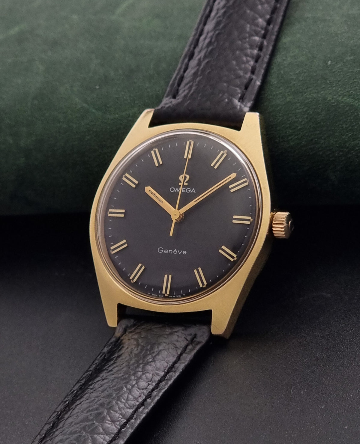 Vintage Omega Geneve 135.041 watch with a gold-tone case, black dial, and black leather strap, resting on a dark textured surface with a green velvet cushion in the background.