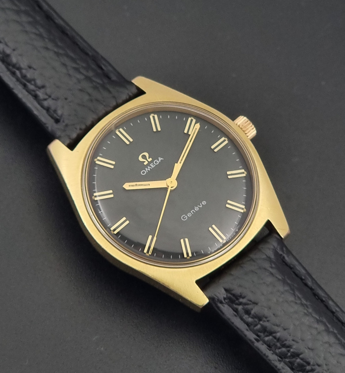 Vintage Omega Geneve 135.041 watch with a gold-tone case, dark gray dial, and black textured leather strap, shown on a dark gray background.