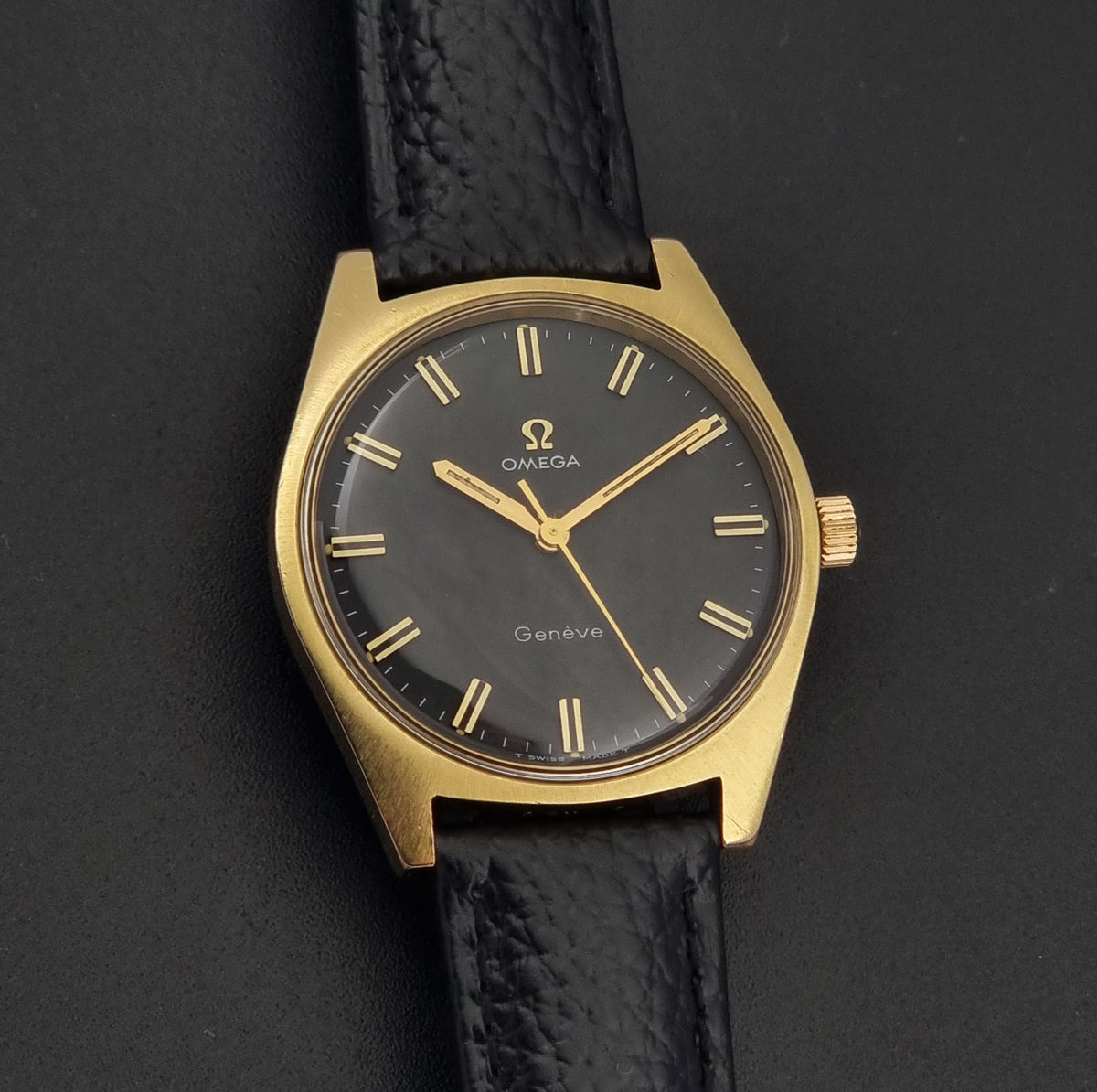 Omega Geneve wristwatch with a gold-tone case, black dial, and black leather strap, photographed on a dark background.