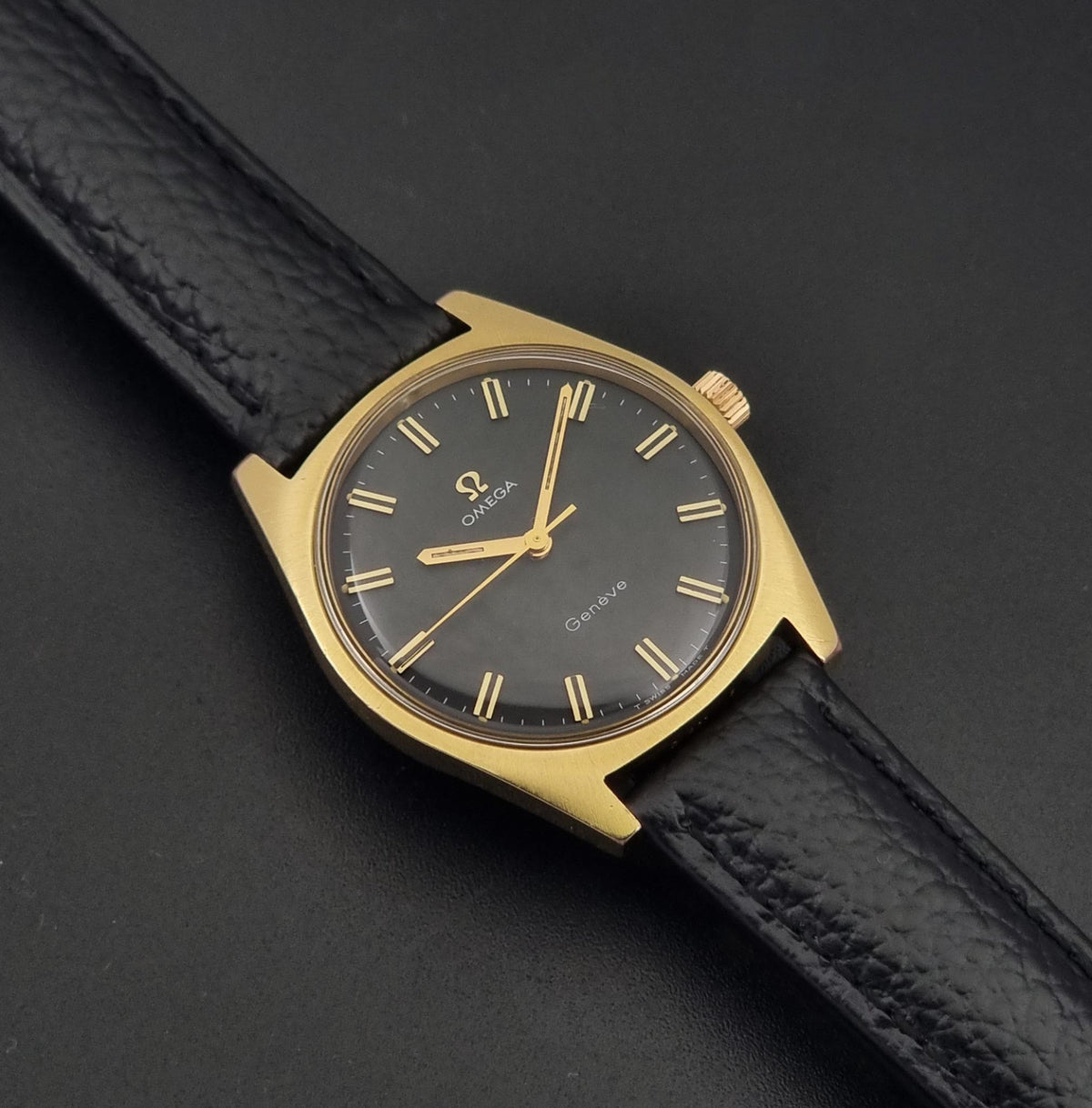 Vintage Omega Geneve 135.041 watch with a gold-tone case, black dial, and black textured leather strap, photographed diagonally on a dark background.