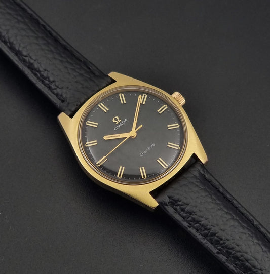 Vintage Omega Geneve 135.041 watch with a gold-tone case, black dial, and black textured leather strap, photographed diagonally on a dark background.
