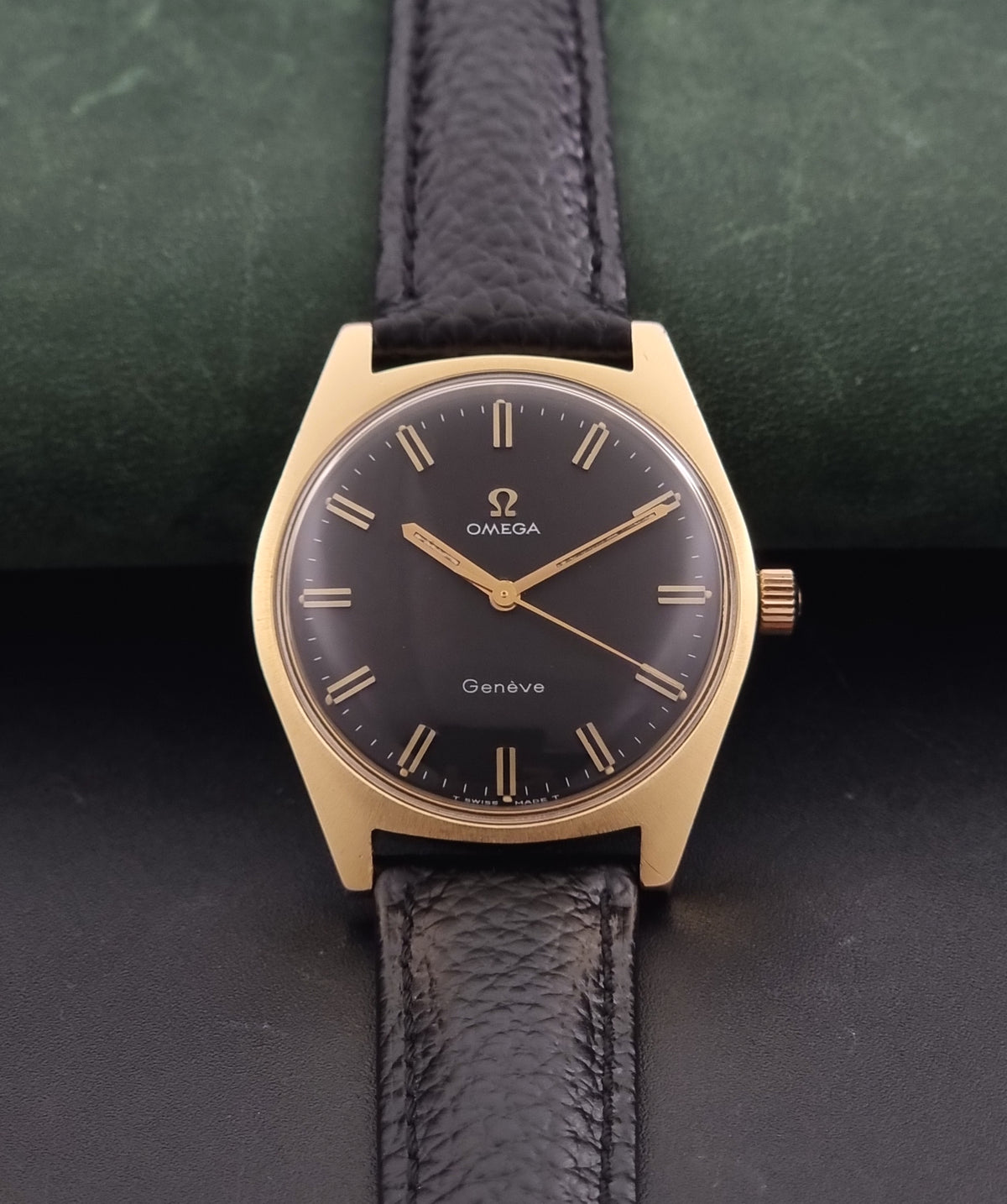 Vintage Omega Geneve 135.041 watch with a gold-tone case, black dial, and textured black leather strap, displayed on a dark green textured background.