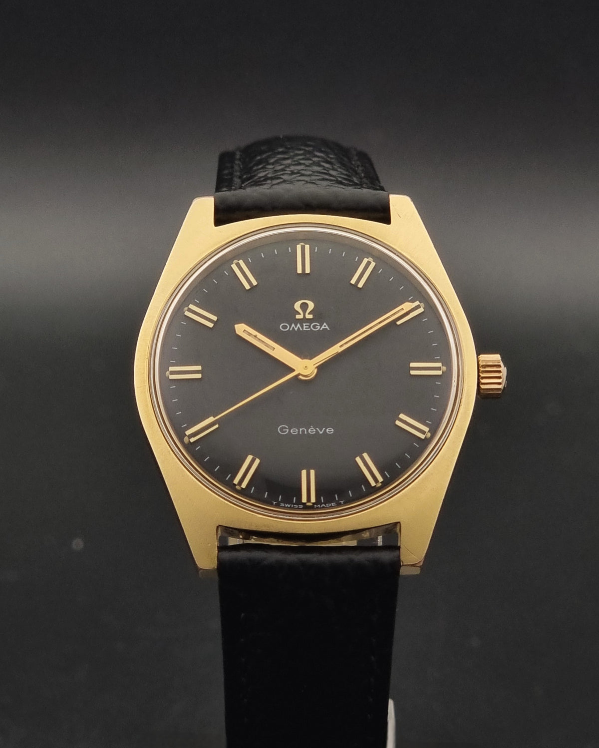 Vintage Omega Geneve 135.041 watch with a gold-tone case, black dial, and black leather strap, shown against a dark gray background.