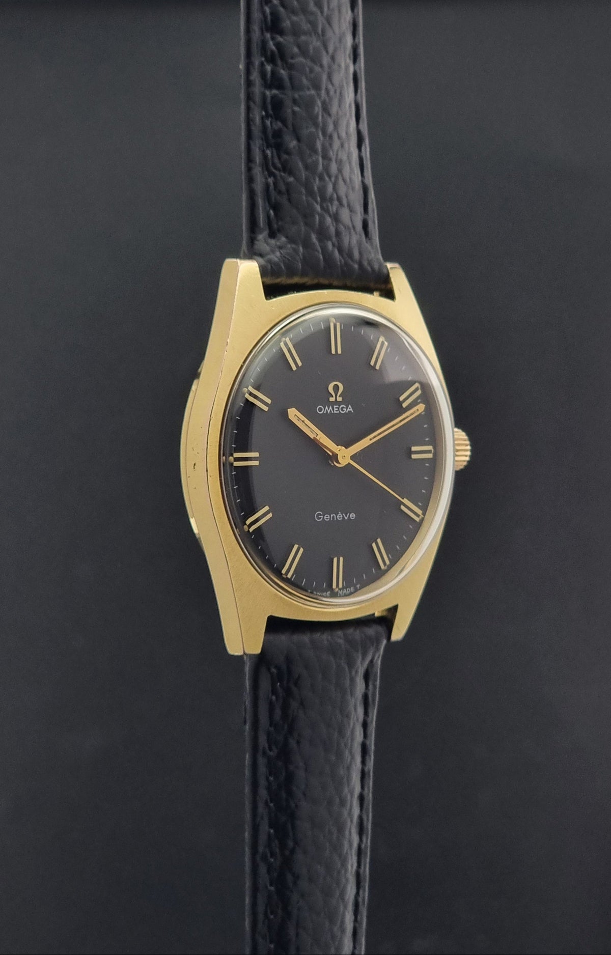 Vintage Omega Geneve 135.041 watch with a gold-tone case, black dial, and black textured leather strap, shown against a dark background.