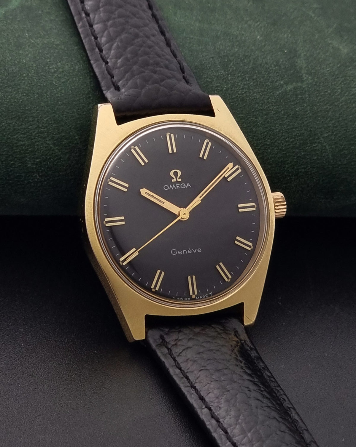 Vintage Omega Geneve 135.041 watch with a gold-tone case, black dial, and black leather strap, resting on a dark textured surface with a green cushion behind it.