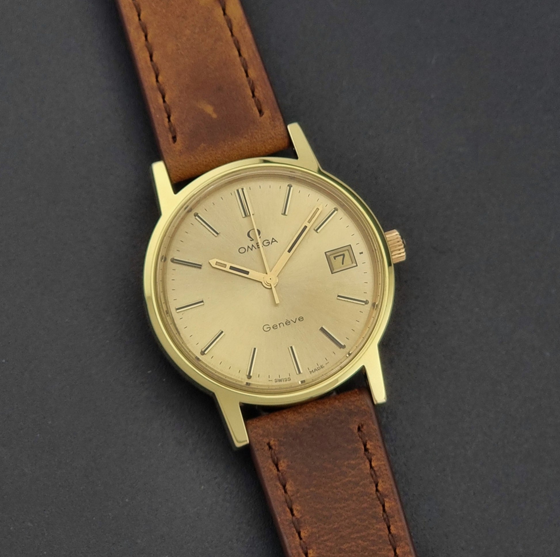 Vintage Omega Wristwatch Geneve 136.0104 with a gold case and brown strap on a dark background front view