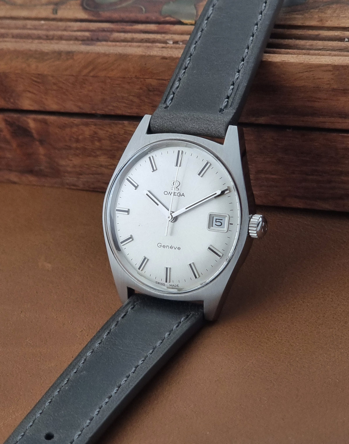 An Omega wristwatch with a silver-toned metal case, white dial, and dark gray leather strap, resting on a brown leather surface with a wooden background.