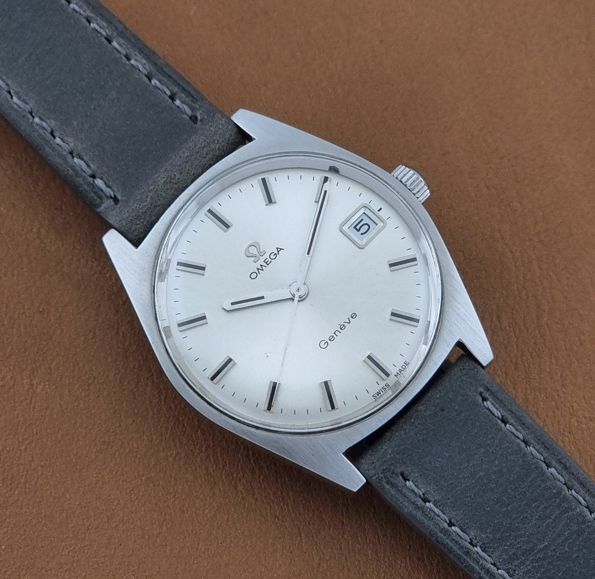 Vintage Omega Geneve 136.041 watch with a silver stainless steel case, white dial, and a dark gray leather strap, shown on a brown textured background.